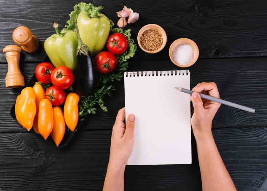 Mão de uma pessoa escrevendo em um bloco de notas em espiral perto de vegetais e temperos em uma superfície de madeira preta.