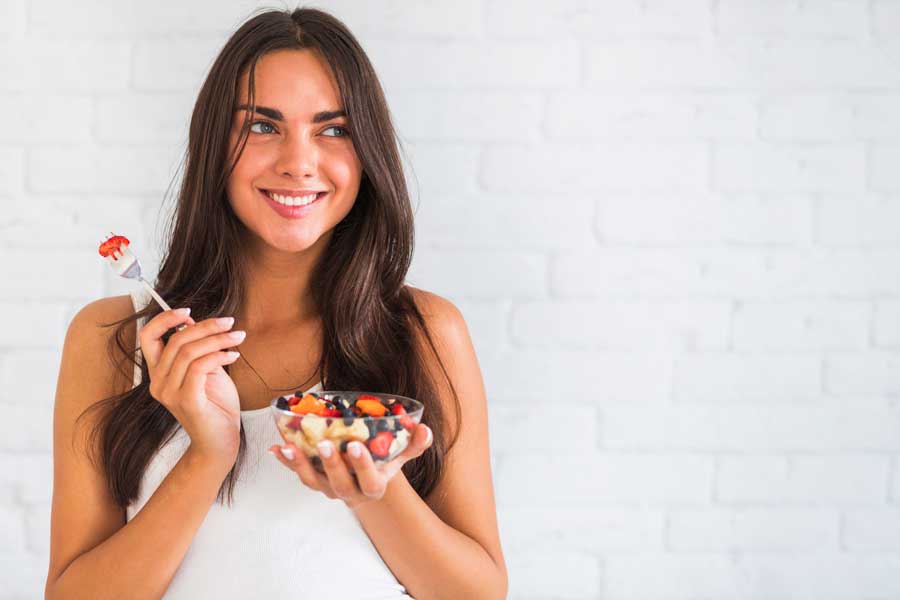 Mulher jovem pensativa e sorridente segurando uma tigela de salada de frutas.