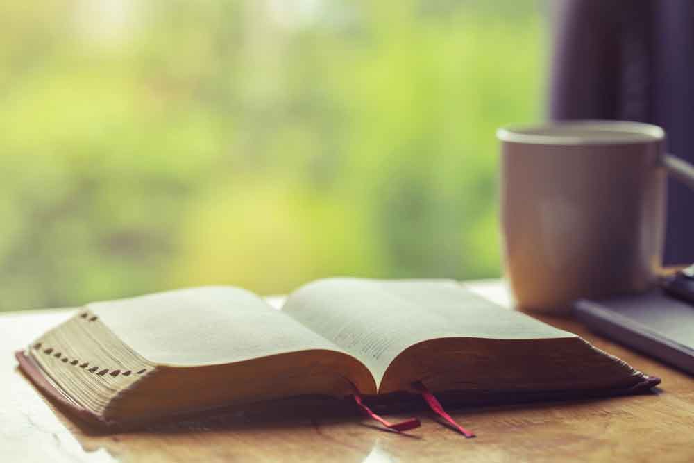 Uma bíblia aberta com uma chávena de café para devoção matinal numa mesa de madeira com luz de janela.