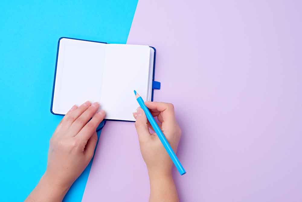 Carnet de journal ouvert et un crayon bleu en bois dans une main féminine droite.