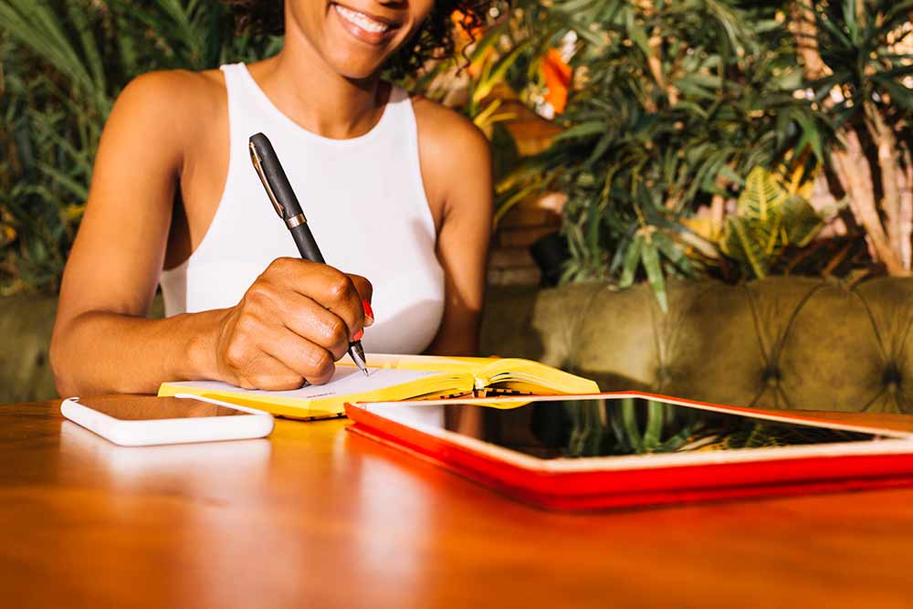 Young woman writing on diary with pen over the wooden table with cellphone and digital tablet.