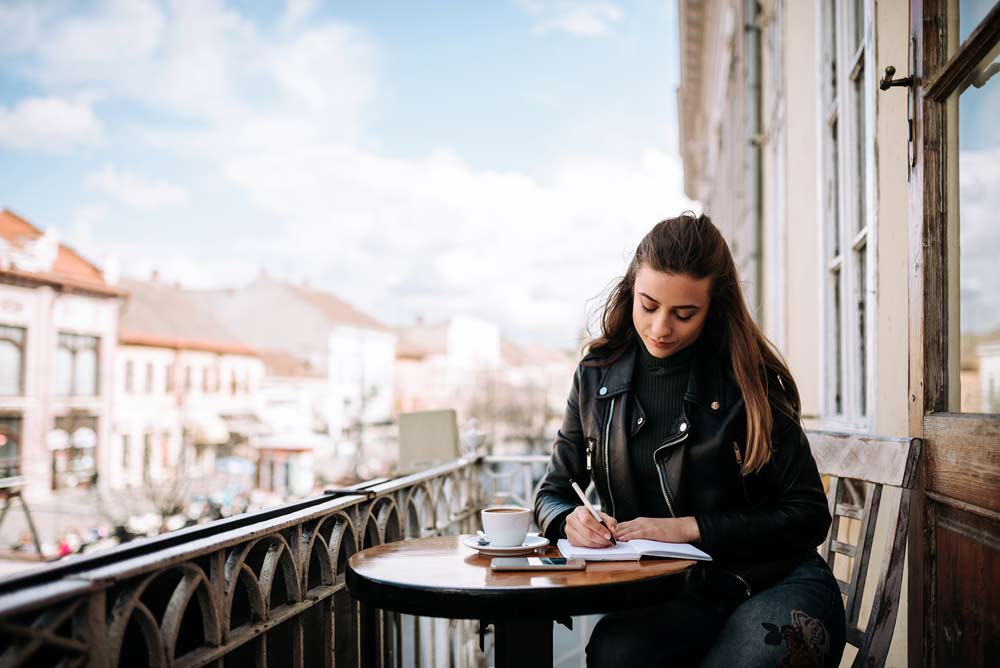Young woman writting in a travel journal diary while sitting on the terrace in the city.