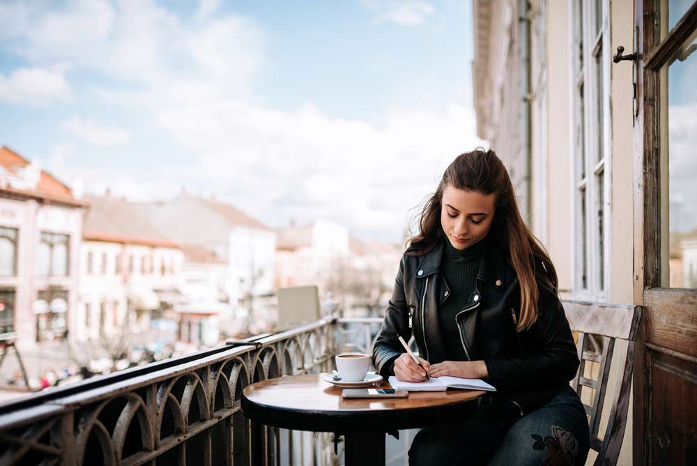 Mujer joven escribiendo en un diario de viaje mientras está sentada en la terraza de la ciudad.